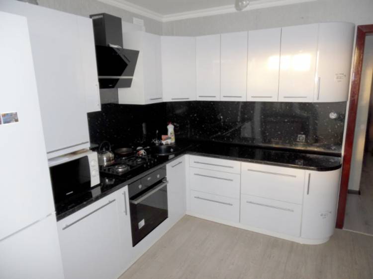 Компактная угловая кухня с глянцевыми белыми фасадами, черной столешницей и фартуком