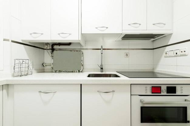 Изображение спереди кухни с белой деревянной мебелью, столешницей, сочетающейся с бытовой техникой, керамической плитой, вытяжкой и белой каменной духовкой под столешницей