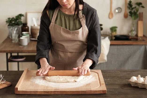 Занятая женщина в фартуке стоит за столом и раскатывает тесто булавкой на деревянной доске на современной кух