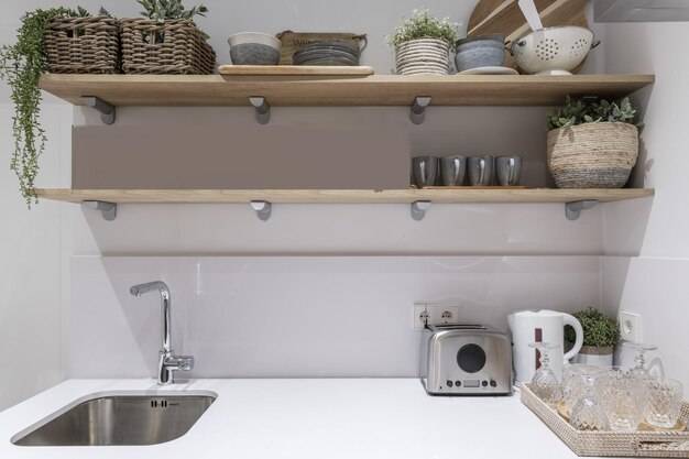 Кухня с белой столешницей с раковиной из нержавеющей стали и деревянными полками цвета экрю с кухонными принадлежностями