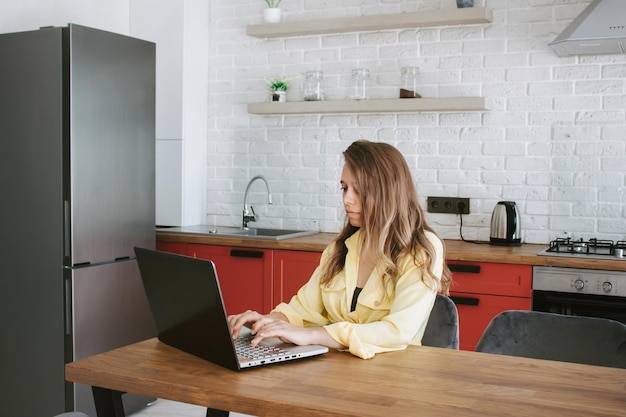 Молодая женщина, сидящая за столом и работающая на ноутбуке из дома на кухне, фрилансер