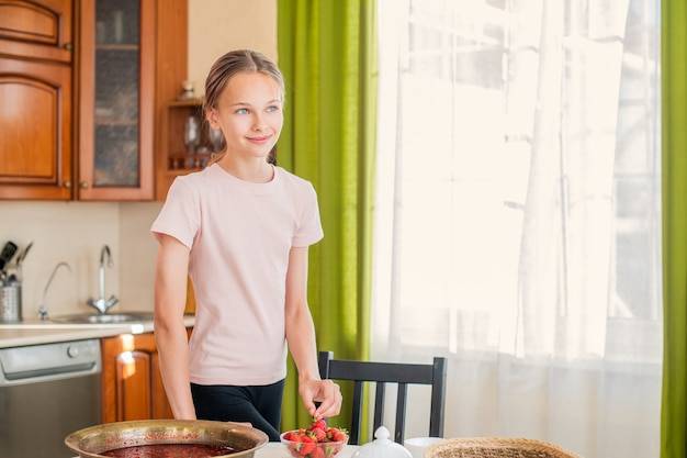 Девушка дома на кухне стоит возле стола, ест клубнику, готовит клубничное варенье, улыбается идеальной улыбкой