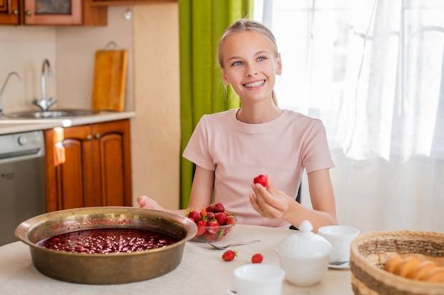 Девушка дома на кухне сидит за столом, ест клубнику, варит клубничное варенье, улыбается безупречной улыбкой