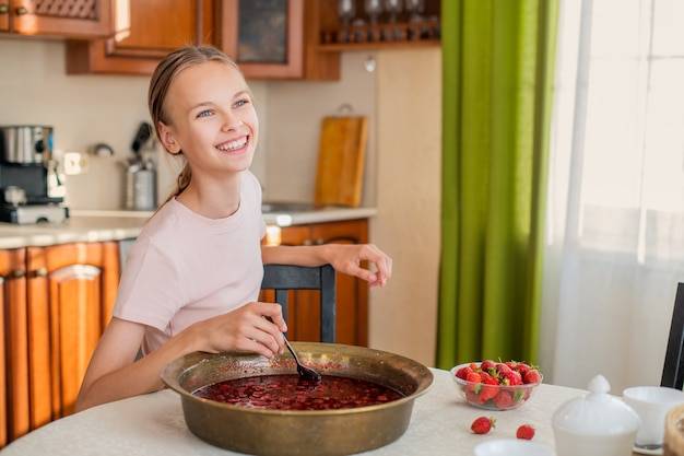 Девушка дома на кухне сидит за столом, ест клубнику, варит клубничное варенье, улыбается безупречной улыбкой